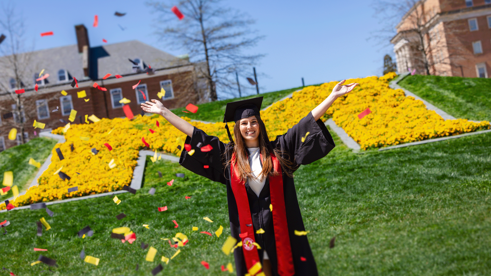 Student in black cap and gown with red stole stands in front of the yellow flower-filled giant M