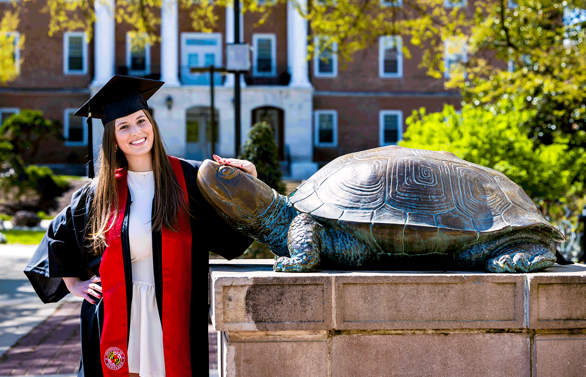 A student in a graduation cap and gown rubs bronze Testudo's nose